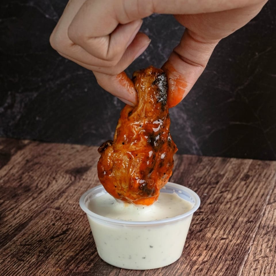 A hand dips a saucy Westside Pizza Express chicken wing into a small cup of ranch dressing.