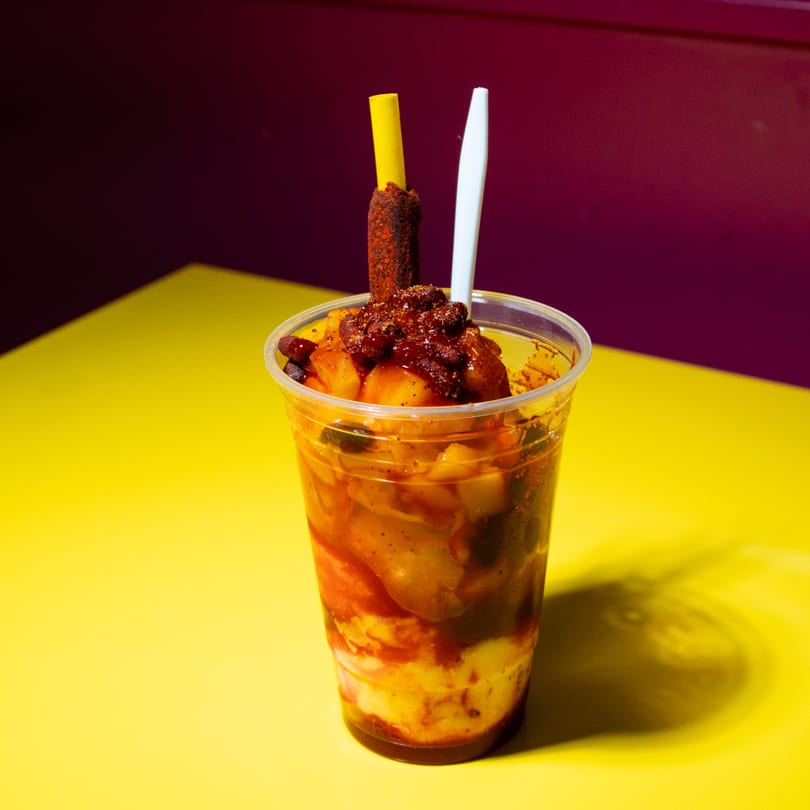 Spicy fruit cup with mango, chili powder, and a tamarind candy stick on a yellow table.