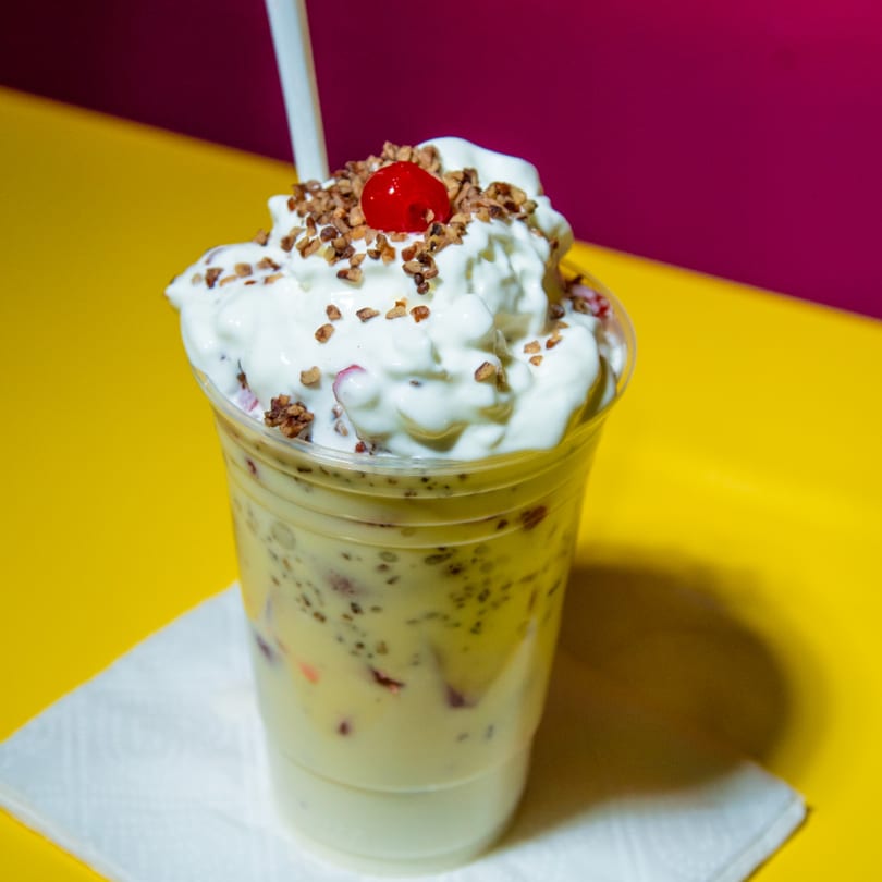 Creamy dessert in a clear cup, topped with whipped cream, chopped nuts, and a bright red cherry.