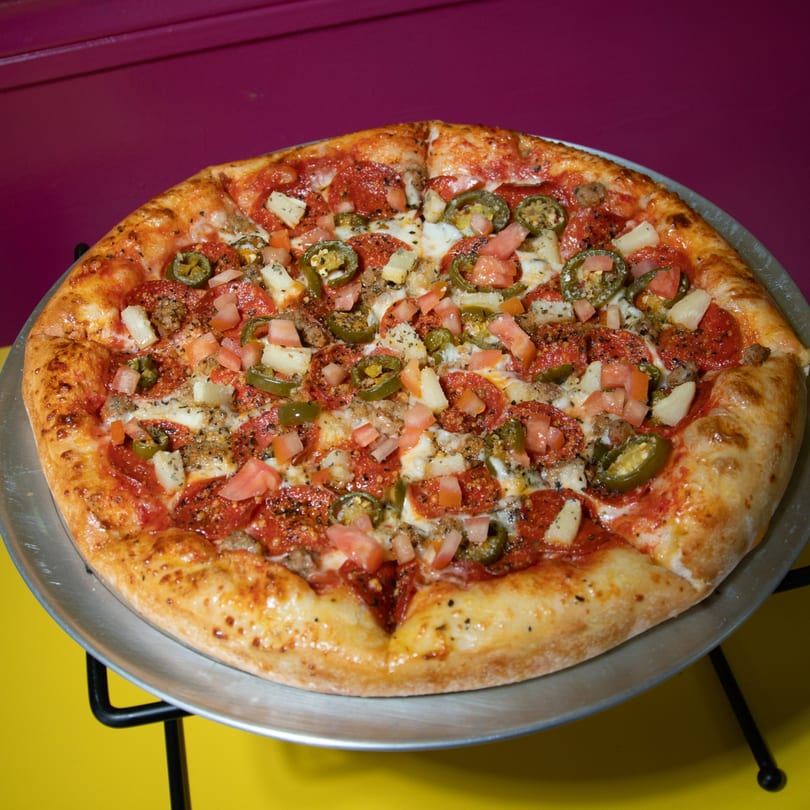Delicious pizza with pepperoni, jalapeños, pineapple, tomatoes, and sausage on a metal tray.